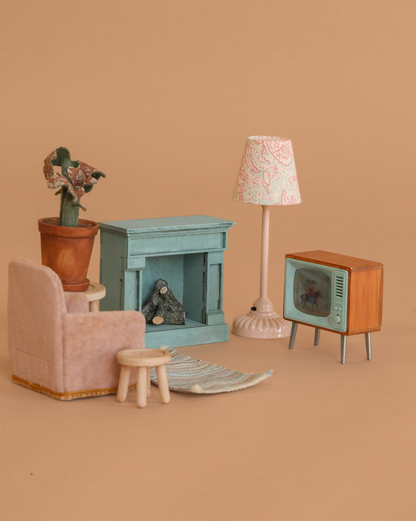 The Maileg Living Room Set features vintage-style mouse furniture: a pink armchair, wooden stool, potted plant, blue fireplace, standing lamp, TV with antennas, and a striped rug—all displayed on a plain beige background.