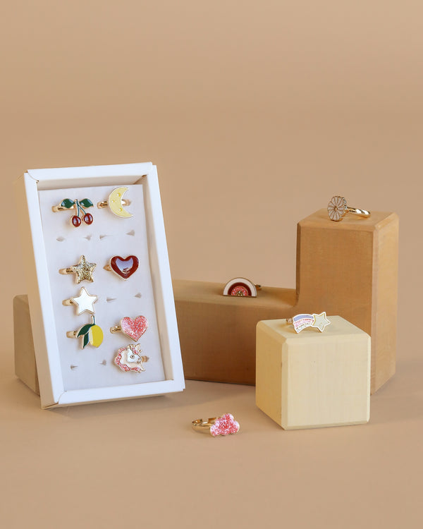 The Konges Sloejd 12 Pack Ring Set features colorful, whimsical adjustable rings with motifs like cherries, moon, stars, heart, bunny, and rainbow—perfect for tiny fingers and arranged on a white card against a neutral background.