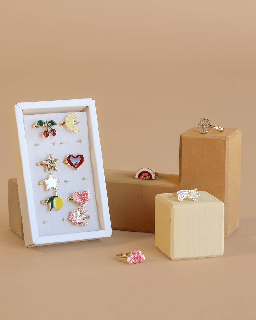 The Konges Sloejd 12 Pack Ring Set features colorful, whimsical adjustable rings with motifs like cherries, moon, stars, heart, bunny, and rainbow—perfect for tiny fingers and arranged on a white card against a neutral background.