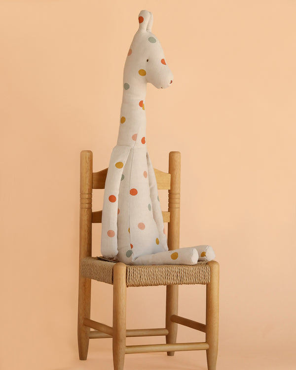 The Maileg Large Giraffe—a charming, soft, and polka-dotted plush safari friend—sits upright on a simple wooden chair against a peach-colored background.
