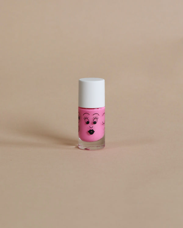 A small pink Nailmatic kids nail polish bottle with a cute face design on a beige background. The cap is white.