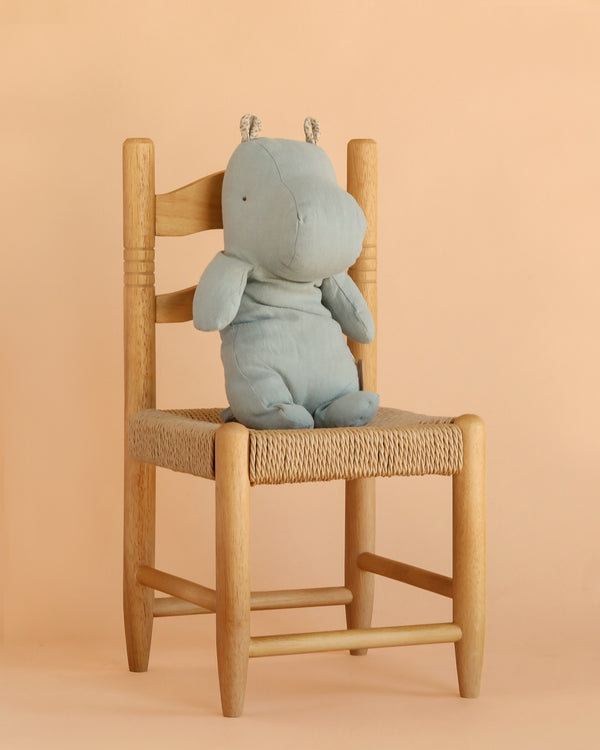 A Maileg Hippo, Medium - Blue Grey, sits on a woven wooden chair against a beige background, ready to join other Safari friends.