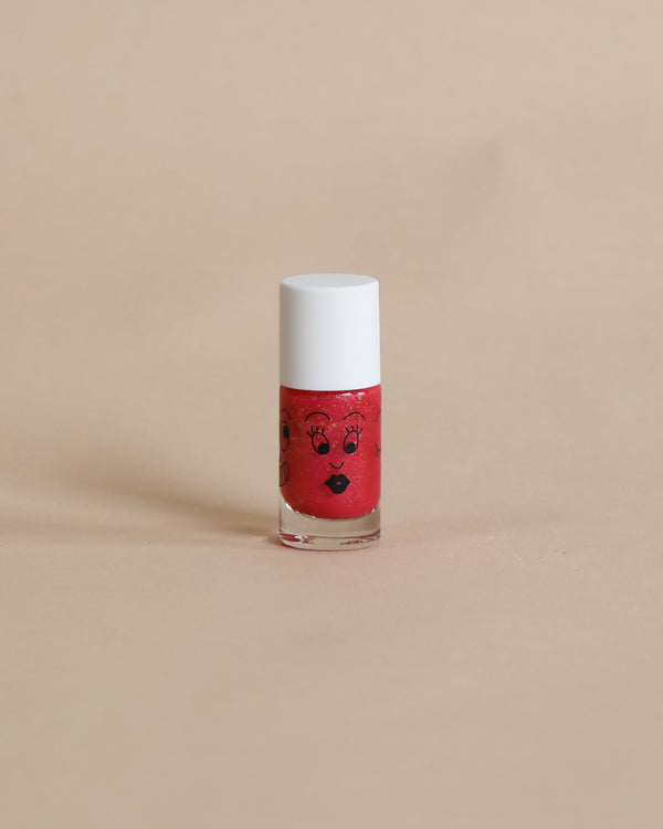 A small bottle of Nailmatic water-based red nail polish with a cartoon face design, featuring eyes and lips, standing upright on a plain beige background.