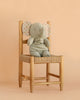 The Maileg Elephant Soft, Medium - Iron Grey, from our Safari friends collection, sits on a woven wooden chair against a plain peach-colored background, showcasing its soft gray fabric and polka-dotted ears.