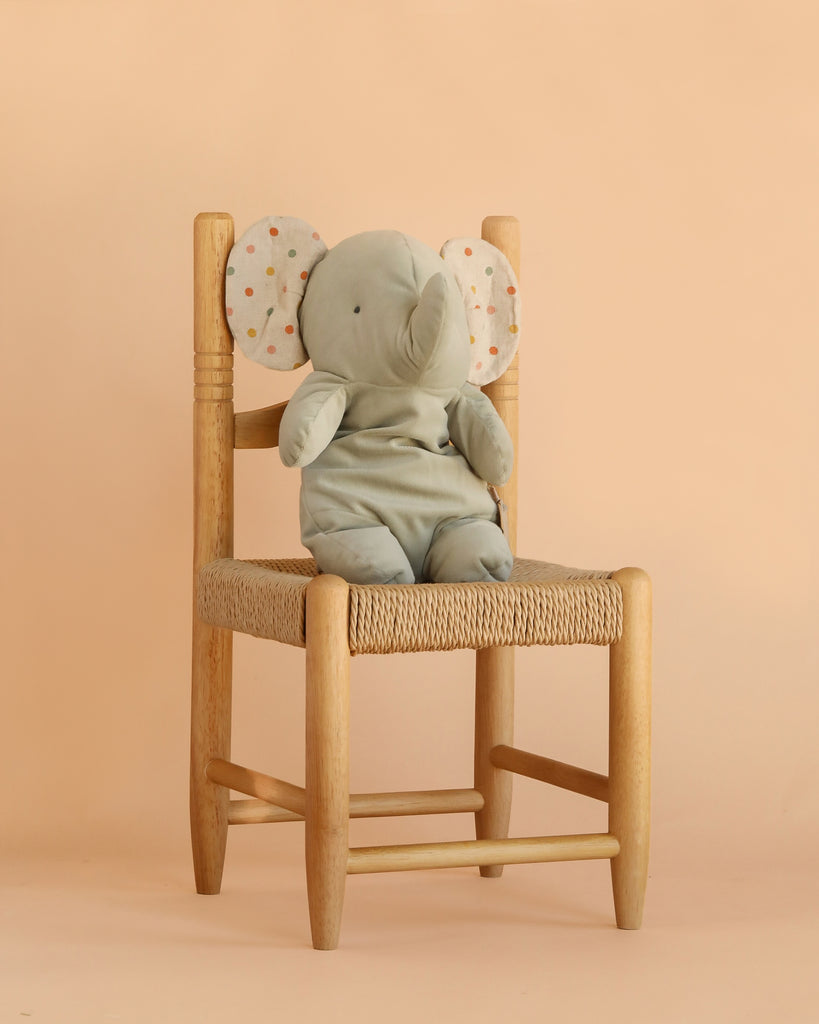 The Maileg Elephant Soft, Medium - Iron Grey, from our Safari friends collection, sits on a woven wooden chair against a plain peach-colored background, showcasing its soft gray fabric and polka-dotted ears.