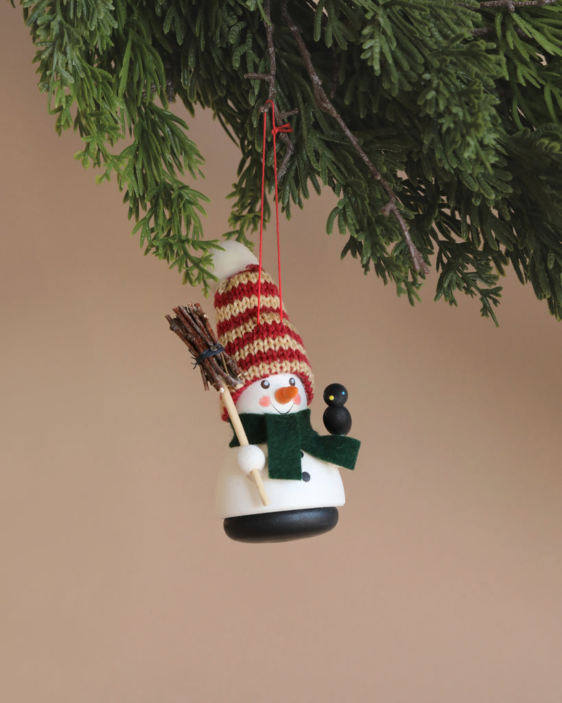 The Christian Ulbricht Ornament - Snowman, featuring a delightful hand-painted snowman adorned with a red and white striped hat, green scarf, and holding a tiny broom, is elegantly suspended from a green branch against a soft beige background.