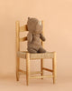 A light brown Maileg Rhino, Medium from the Safari Friends collection sits upright on a light wooden chair with a woven seat, crafted from soft fabric, against a beige background.