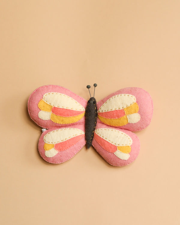 A colorful handcrafted felt butterfly with pink, yellow, and orange layered wings, displayed on a light beige background, crafted as ethically made handcrafted nursery décor.