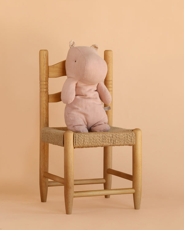 The Maileg Hippo, Medium - Light Rose, sits upright on a wooden chair with a woven seat, set against a light beige background.