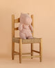 The Maileg Hippo, Medium - Light Rose, sits upright on a wooden chair with a woven seat, set against a light beige background.