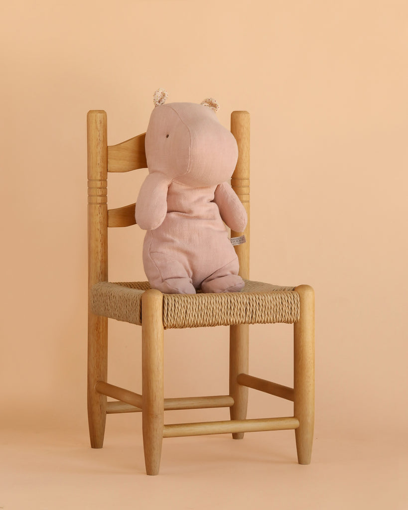 The Maileg Hippo, Medium - Light Rose, sits upright on a wooden chair with a woven seat, set against a light beige background.