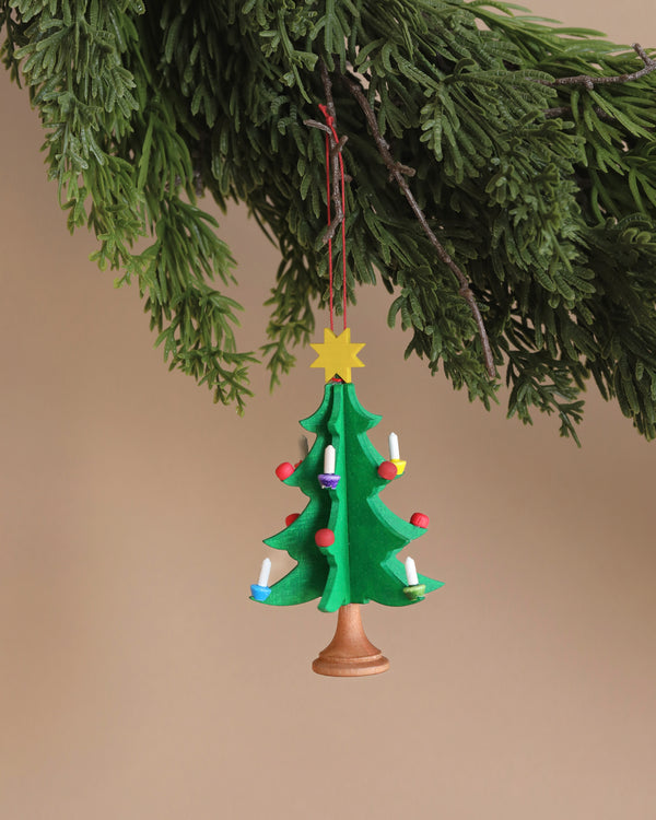 Against a plain background, the charm of a Christian Ulbricht Ornament - Christmas Tree is showcased as it hangs from a branch; this small, colorful hand-painted wooden piece features candles and a yellow star at the top.