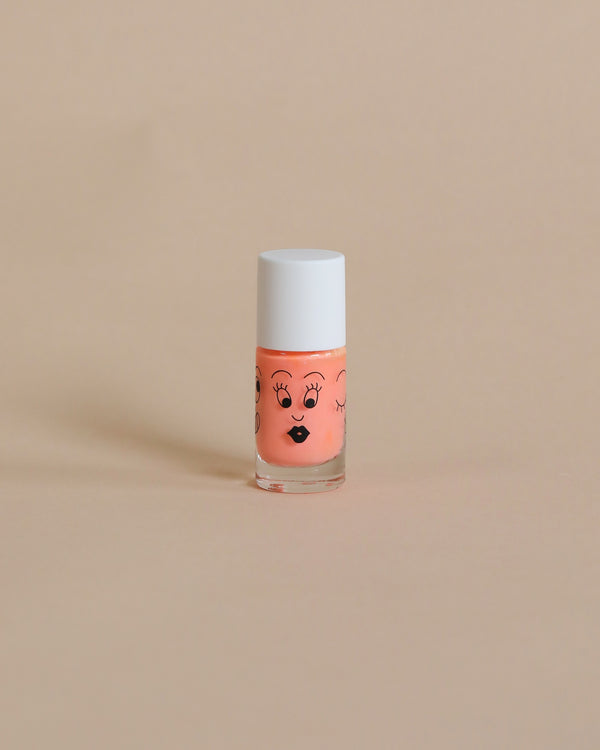 A small bottle of peach-colored Nailmatic washable nail polish with a cute face drawn on it, featuring eyes, eyebrows, and lips, set against a plain beige background.
