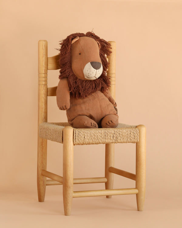 The Maileg Lion Stuffed Animal, Medium, with its shaggy mane and charming handmade details, sits upright on a wooden chair with a woven seat against a beige background—sure to become one of your new favorite safari friends.
