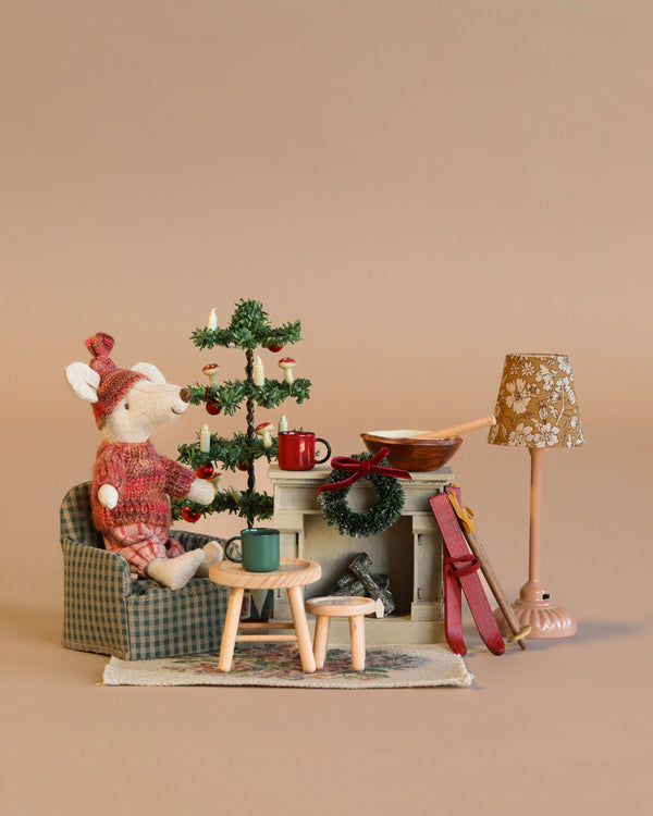 A small Maileg Winter Ski Mouse in a red sweater sits by a miniature tree, Maileg Mouse Fireplace, and table with mugs, creating a festive scene with the Maileg Cozy Christmas Set.