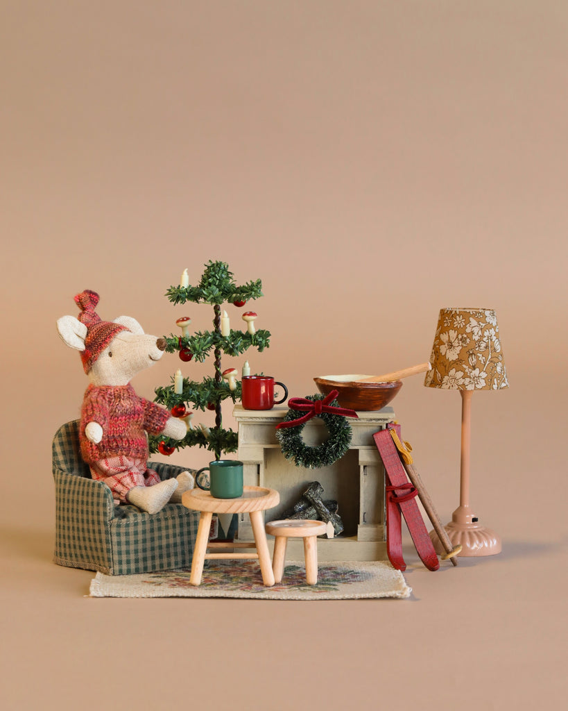 A small Maileg Winter Ski Mouse in a red sweater sits by a miniature tree, Maileg Mouse Fireplace, and table with mugs, creating a festive scene with the Maileg Cozy Christmas Set.