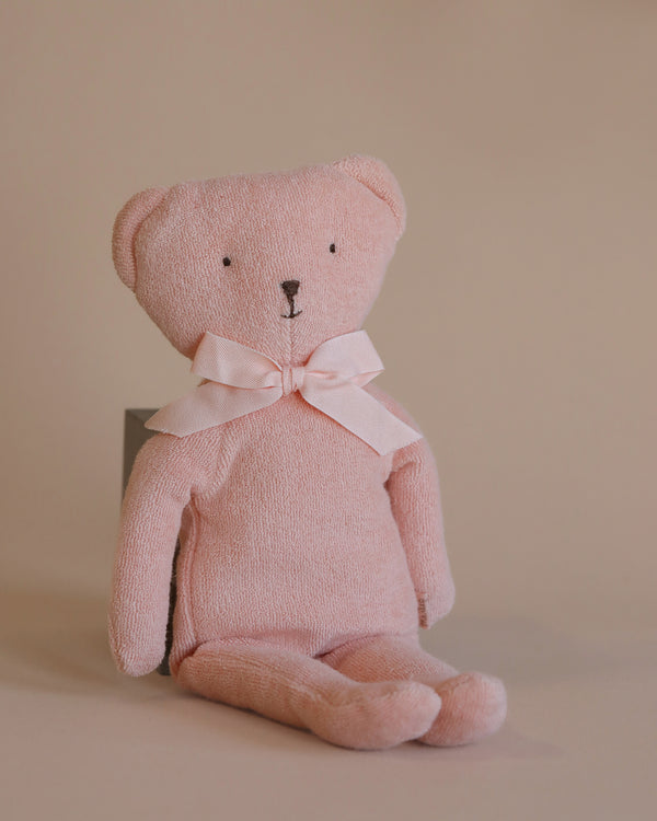 The Maileg Teddy - Rose, in a gentle pink hue with a matching ribbon around its neck, is seated against a neutral beige background. Made from plush cotton, this teddy bear features simple stitched details and boasts a cozy, soft fabric texture.