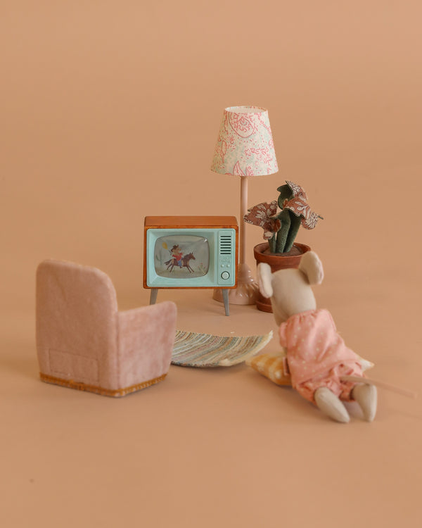 A Maileg Cozy TV Time Set features a Big Sister Mouse in a pink dress relaxing on a Miniature Rug, with a pink armchair, standing lamp, and potted plant in a cozy room against a warm beige background.