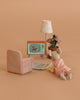 A Maileg Cozy TV Time Set features a Big Sister Mouse in a pink dress relaxing on a Miniature Rug, with a pink armchair, standing lamp, and potted plant in a cozy room against a warm beige background.