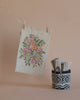 The Maileg Flower Rug, Medium, featuring a floral embroidery on beige fabric, hangs gracefully from a clothesline with wooden pegs. Next to it sits a charming black and white patterned miniature basket, reminiscent of those in cozy dollhouses, cradling folded striped fabrics. The backdrop is a simple yet warm beige hue.