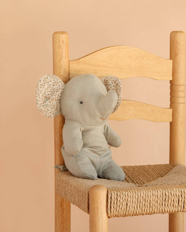 The Maileg Elephant, Small - Iron Grey from the Safari Friends collection, crafted with the softest fabric and adorned with patterned ears, sits on a woven wooden chair against a beige background.