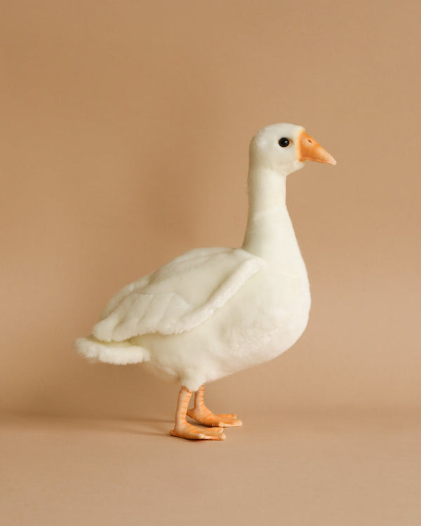 A Young Duck Stuffed Animal with white fur, an orange beak, and matching orange feet is standing against a beige background. The toy appears soft and cuddly, with realistic features that mimic a real duck.