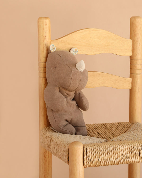 The Maileg Rhino, Small - Chocolate, crafted from soft fabric in a rich brown hue, sits on a woven wooden chair against a beige background—ready to join your collection of Safari friends.