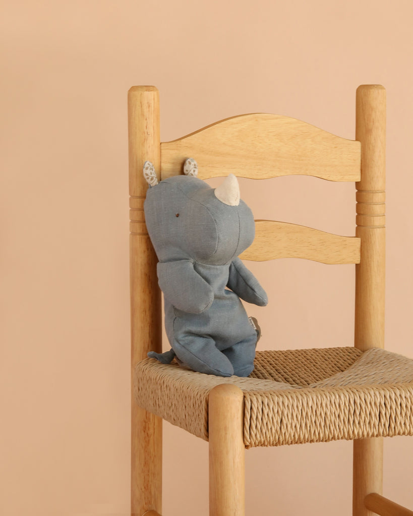 The Maileg Rhino, Small - Dusty Blue, one of our adorable Safari friends, sits upright on a wooden chair with a woven seat against a plain beige background.