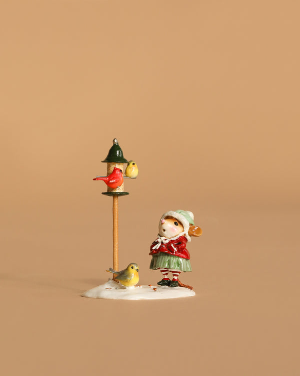 The Wee Forest Folk Tiny Collectible - Feathered Friends depicts a girl in a red coat and green skirt standing by a bird feeder with birds, set on snowy ground against a plain beige background.