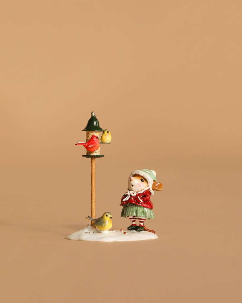 The Wee Forest Folk Tiny Collectible - Feathered Friends depicts a girl in a red coat and green skirt standing by a bird feeder with birds, set on snowy ground against a plain beige background.