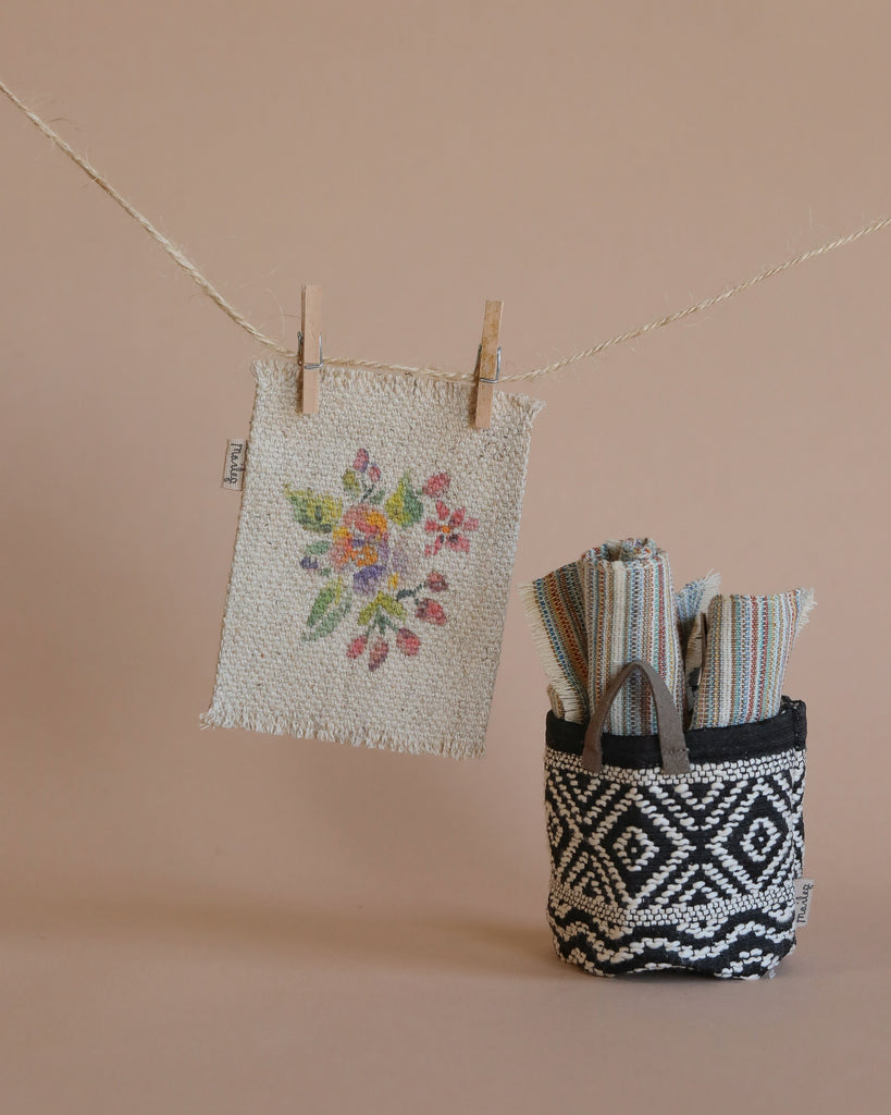 A Maileg Flower Rug, Small, with a floral pattern is hanging on a clothesline with wooden clips. Beside it, a black and white patterned basket holds folded fabrics reminiscent of textiles for doll houses. The background features a solid light brown color.