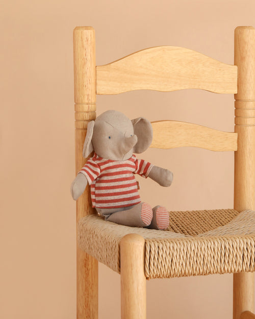 Maileg Little Jumbo the Elephant, a small linen stuffed animal wearing a red and white striped shirt, sits on a woven seat wooden chair against a beige background, making this plush toy a charming accent for any space.