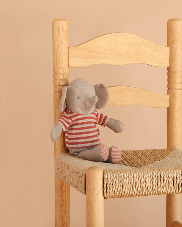 Maileg Little Jumbo the Elephant, a small linen stuffed animal wearing a red and white striped shirt, sits on a woven seat wooden chair against a beige background, making this plush toy a charming accent for any space.