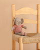 Maileg Little Jumbo the Elephant, a small linen stuffed animal wearing a red and white striped shirt, sits on a woven seat wooden chair against a beige background, making this plush toy a charming accent for any space.
