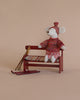 A small stuffed mouse from the Maileg Mouse Bench - Red collection wears a red knit sweater and hat while seated on a wooden bench, accompanied by tiny, hand-painted skis and poles against a beige background.