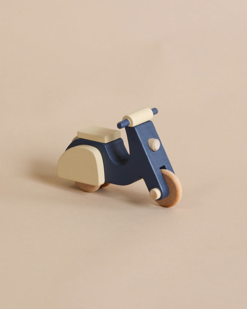 A small, wooden toy scooter with a minimalist design, crafted from responsibly sourced beech wood. The Wooden Scooter is navy blue with cream-colored accents on the seat, handlebars, and rear sections, as well as light wooden wheels coated in nontoxic paint. The background is a plain, beige surface.