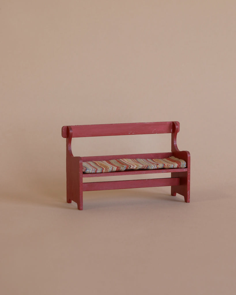 The Maileg Mouse Bench - Red, featuring a vibrant striped cushion and hand-painted in small red details, is pictured against a simple beige backdrop, as if anticipating the arrival of an adorable mouse family to claim it as their home. (Ships in approximately one week.)