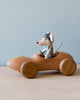 A small stuffed mouse toy, wearing a blue cap and vest, sits in a Handmade Wooden Race Car with large, round wheels crafted from handmade beech wood. The car boasts an ultra smooth finish thanks to natural linseed oil. Set against a light blue background, it rests on a smooth, light-colored wooden surface.