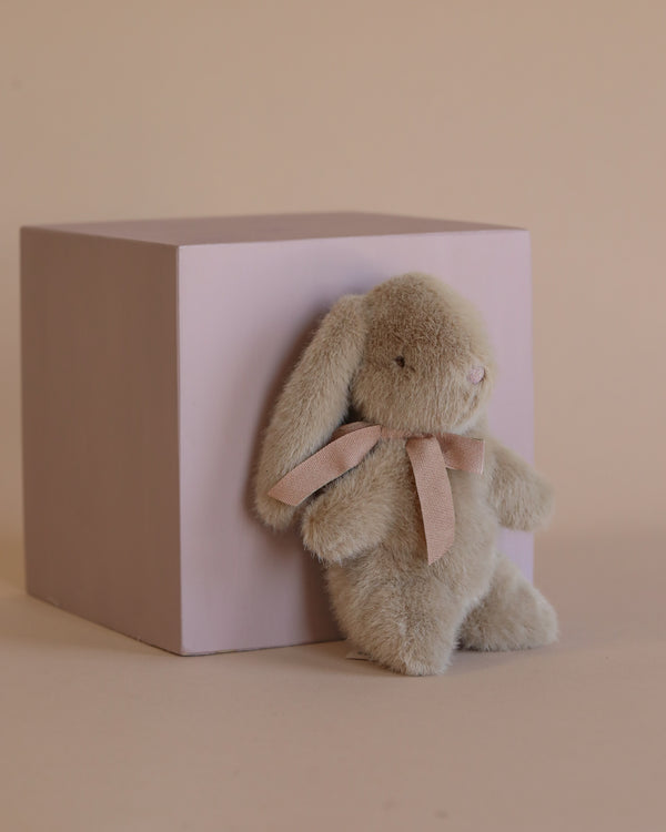 The Maileg Mini Plush Bunny - Dusty Brown, complete with polyester fillings and adorned with a pink ribbon around its neck, leans against a light pink cube set against a beige background.