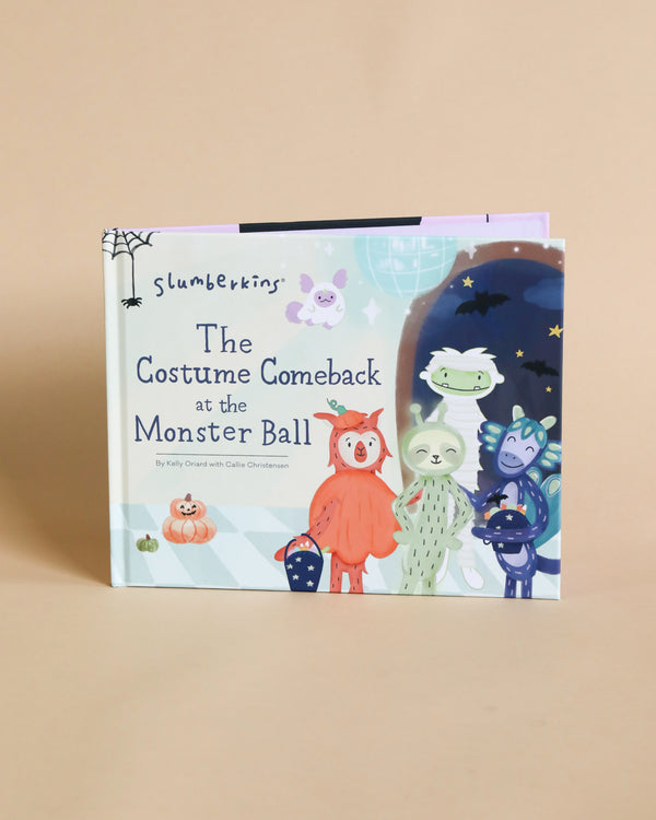 A colorful children's book titled "Slumberkins "The Costume Comeback" Halloween Hardcover Book", displayed upright against a light beige background. The cover features cute cartoon monsters in costumes, emphasizing self.