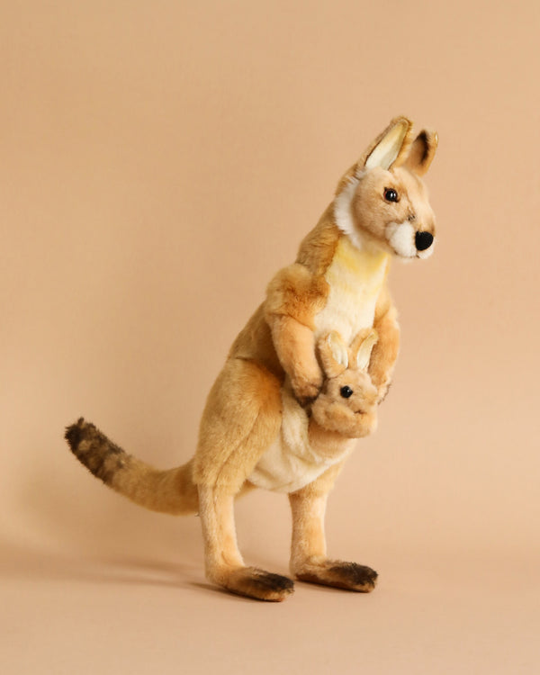 A Kangaroo Mom Stuffed Animal with a joey in its pouch stands against a plain beige background. The hand-sewn kangaroo has a light brown body, dark brown paws, and a lighter underbelly. The joey peeking out from the pouch matches the colors of the adult kangaroo, showcasing realistic features typical of HANSA animals.