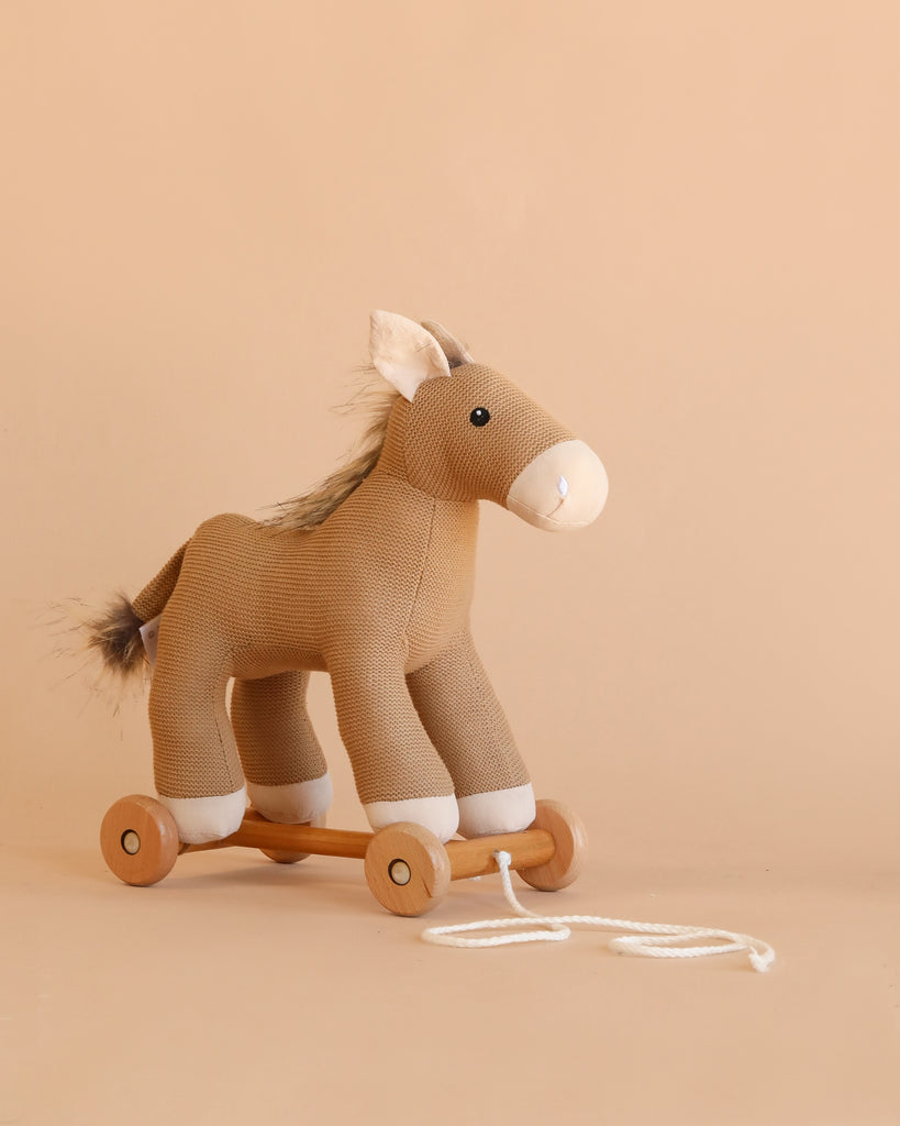 Pull-Along Henry is a brown plush wooden horse toy with white accents, standing on four wooden wheels and attached to a pull string. Set against a plain beige background, this charming pull-along toy helps develop toddler motor skills.