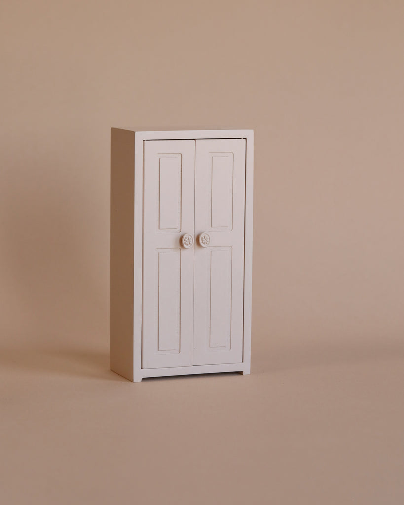 A small, minimalist light rose Maileg Mouse Closet stands against a plain beige background. Inside, golden hangers add a touch of elegance behind its two doors with simple round knobs, creating a clean and uncluttered look.