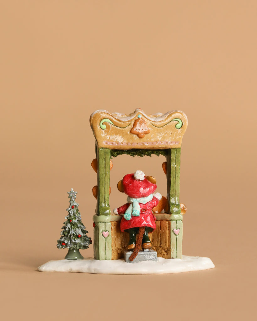 The Wee Forest Folk Tiny Collectible - A Gingerbread Christmas features a girl in a red coat and hat at a decorated booth beside a snow-covered Christmas tree, set against a plain brown background.