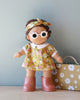 A handmade Dinkum Doll - Sprout (Extended Pack) stands in front of a light blue background, wearing a yellow floral dress, white-collared shirt, white-rimmed glasses, a matching floral headband, and brown boots. The soft cotton doll features exquisite embroidered details. Next to the doll is a beige suitcase adorned with white flowers.