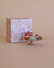 The Maileg Miniature Cakes & Cakestand showcases an exquisite selection of colorful petit fours, including a brown square cake adorned with a red berry. In the backdrop, a decorative box embellished with floral patterns and the text "petit fours" beautifully contrasts against a beige background.