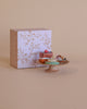A small cake stand displays assorted colorful treats from the Maileg Pastry Chef Set With Grapat Wooden Bake Set in front of a decorative pastel "petit four" box on a beige background.