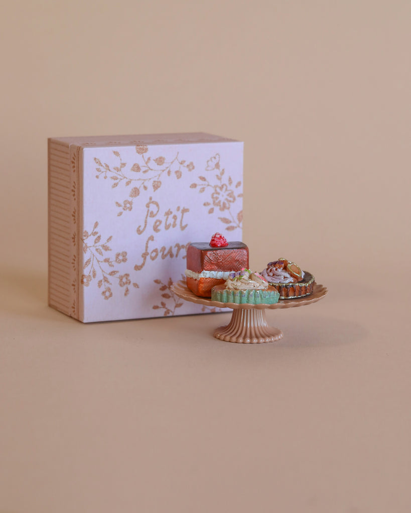 A small cake stand displays assorted colorful treats from the Maileg Pastry Chef Set With Grapat Wooden Bake Set in front of a decorative pastel "petit four" box on a beige background.