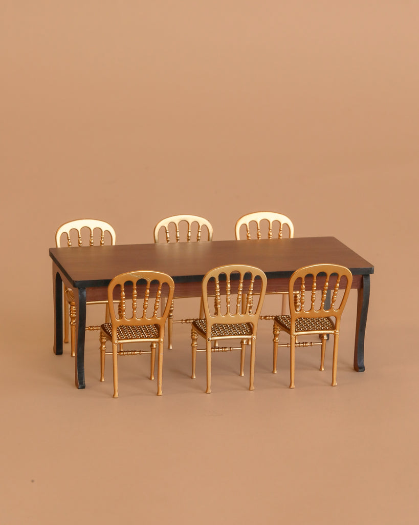The Maileg Dining Room Set features a miniature dark brown long dining table with six gold mouse-size chairs, all arranged against a plain beige background.