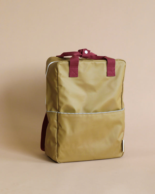 A shiny, golden Sticky Lemon Backpack | Uni | Inventor Green with burgundy straps stands against a light tan backdrop. It features a minimalist design with a reflective horizontal stripe and utilizes waterproof nylon.